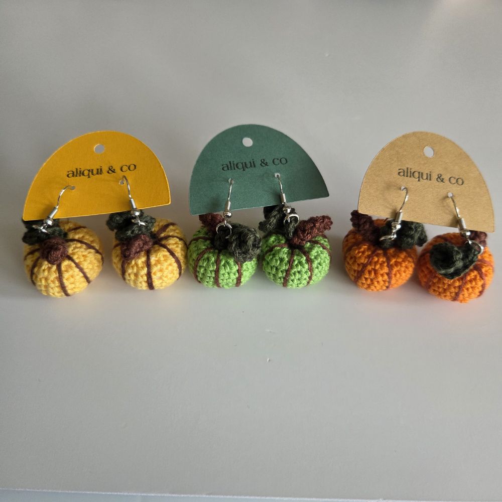 Three pairs of crocheted pumpkin drop earrings on display cards. The left pair has yellow and brown pumpkins on a yellow card, the middle pair has green and brown pumpkins on a green card, and the right pair has orange and brown pumpkins on a tan card.