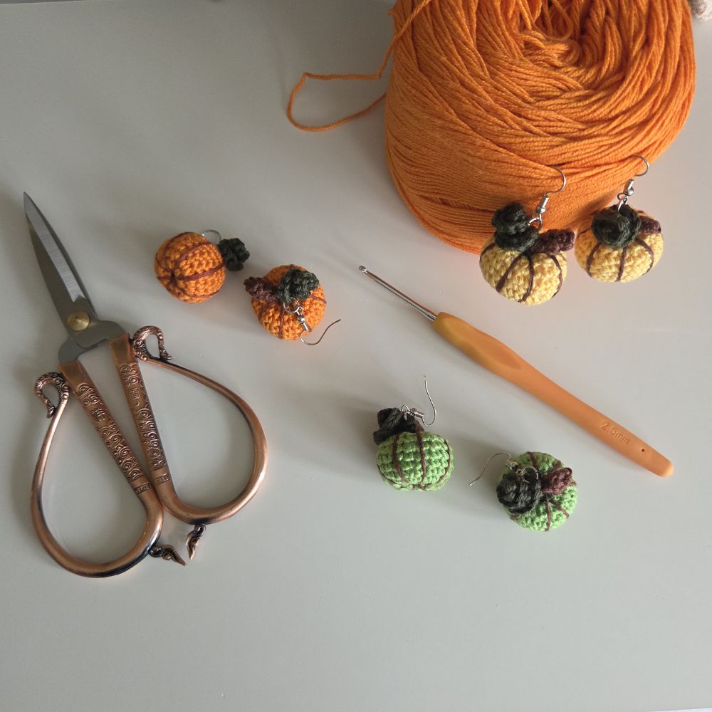 A flat-lay of crochet hook earrings shown with crafting tools and yarn on a light gray surface. The set includes two green and two orange mini-pumpkin-shaped earrings with black stems, a large ball of orange yarn, a wooden-handled crochet hook, and copper scissors.