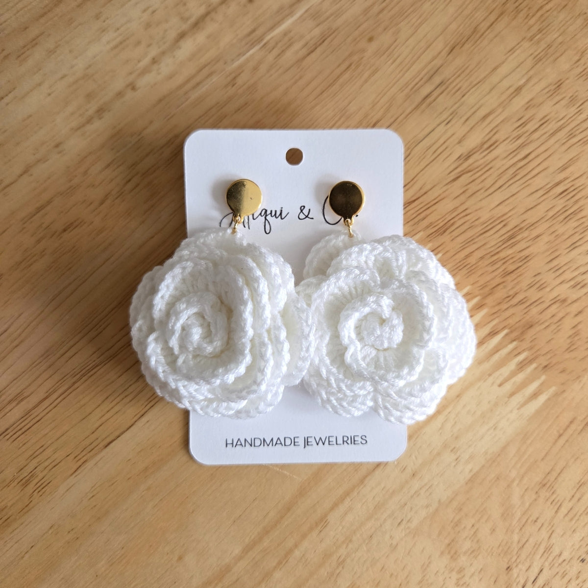 White floral crochet rose earrings on a wooden surface with a branded card.
