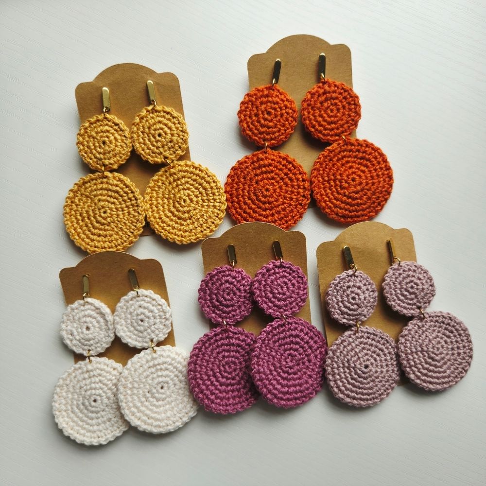 Four pairs of handmade crocheted dangle earrings displayed on brown cardboard hangers against a light gray background. The earrings feature circular spiral patterns in yellow-gold, orange, white, and magenta-pink.