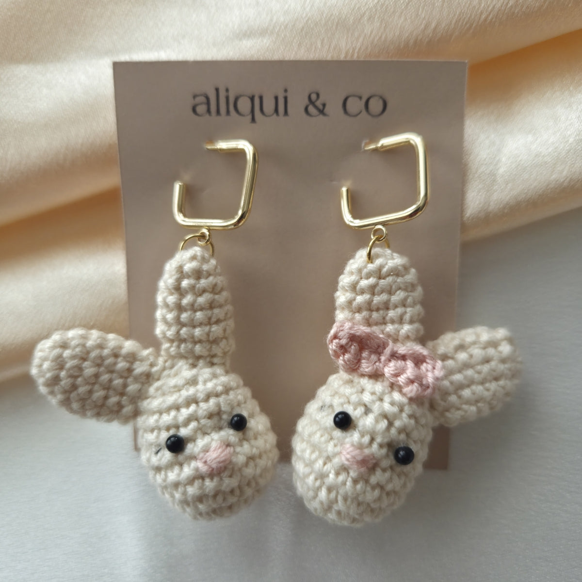 Crocheted bunny earrings with a pink bow on a 'aliqui & co' card.