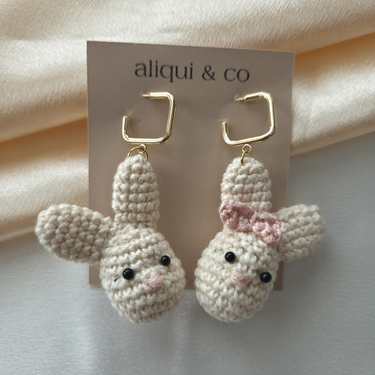 Pair of crocheted bunny earrings on a 'aliqui & co' card against a beige fabric background.