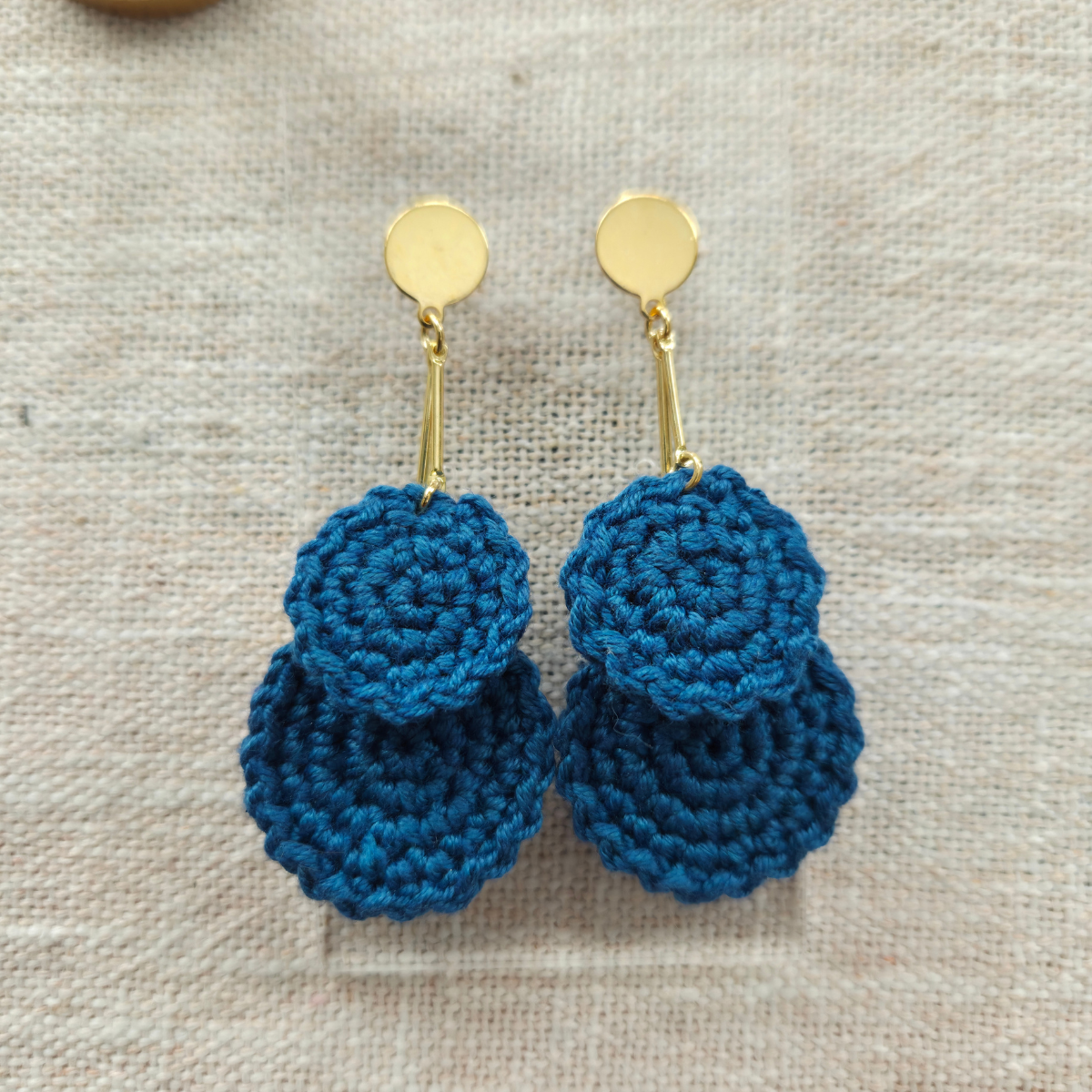 Blue crocheted earrings with gold hooks on a beige fabric background