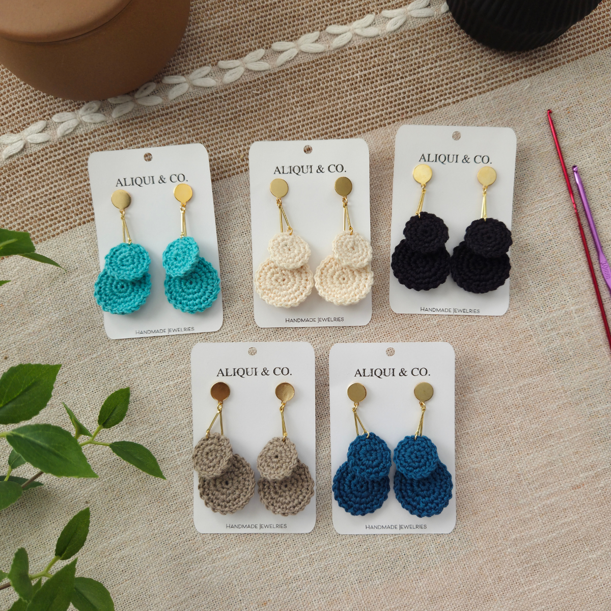 Set of crocheted earrings on Aliqui & Co cards on a textured surface with crochet hooks.