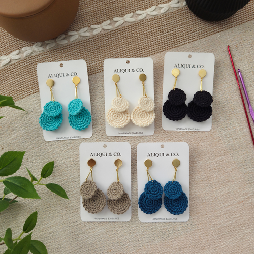 Set of crocheted earrings on Aliqui & Co cards on a textured surface with crochet hooks.