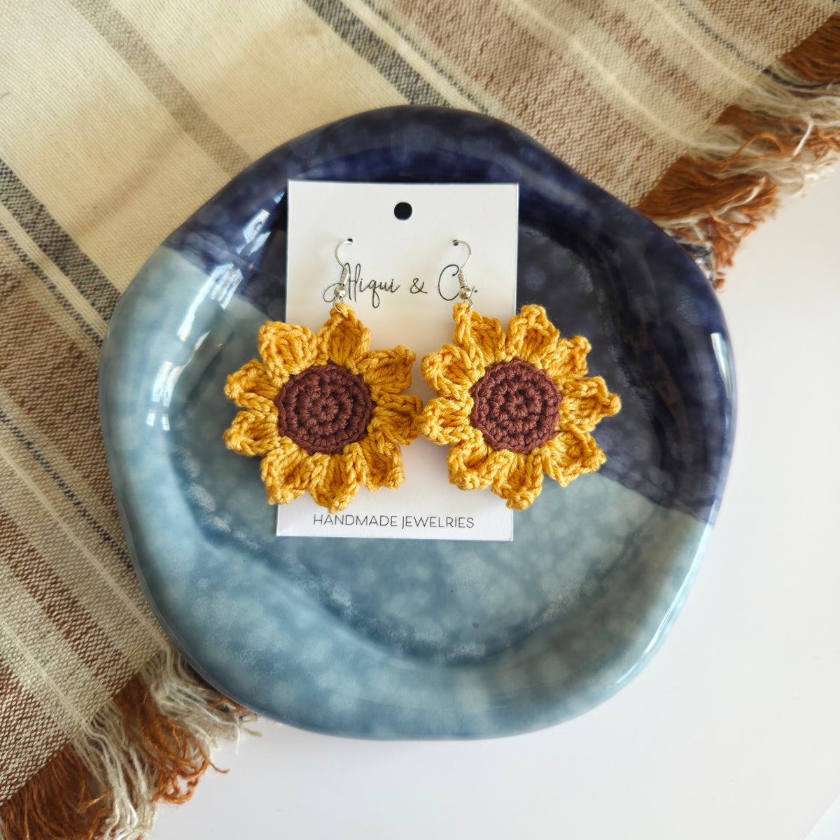Crocheted sunflower earrings on a blue dish with a brand card.