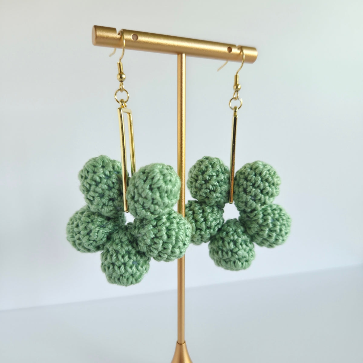 sage green crocheted earrings on a gold stand against a light background