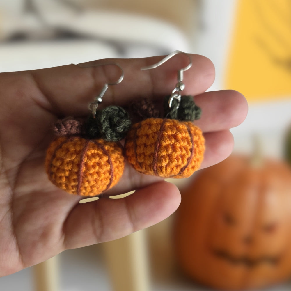 A hand holds a pair of orange crocheted pumpkin earrings with dark green and brown stems and silver fishhook wires. A blurred jack-o'-lantern sits in the background.