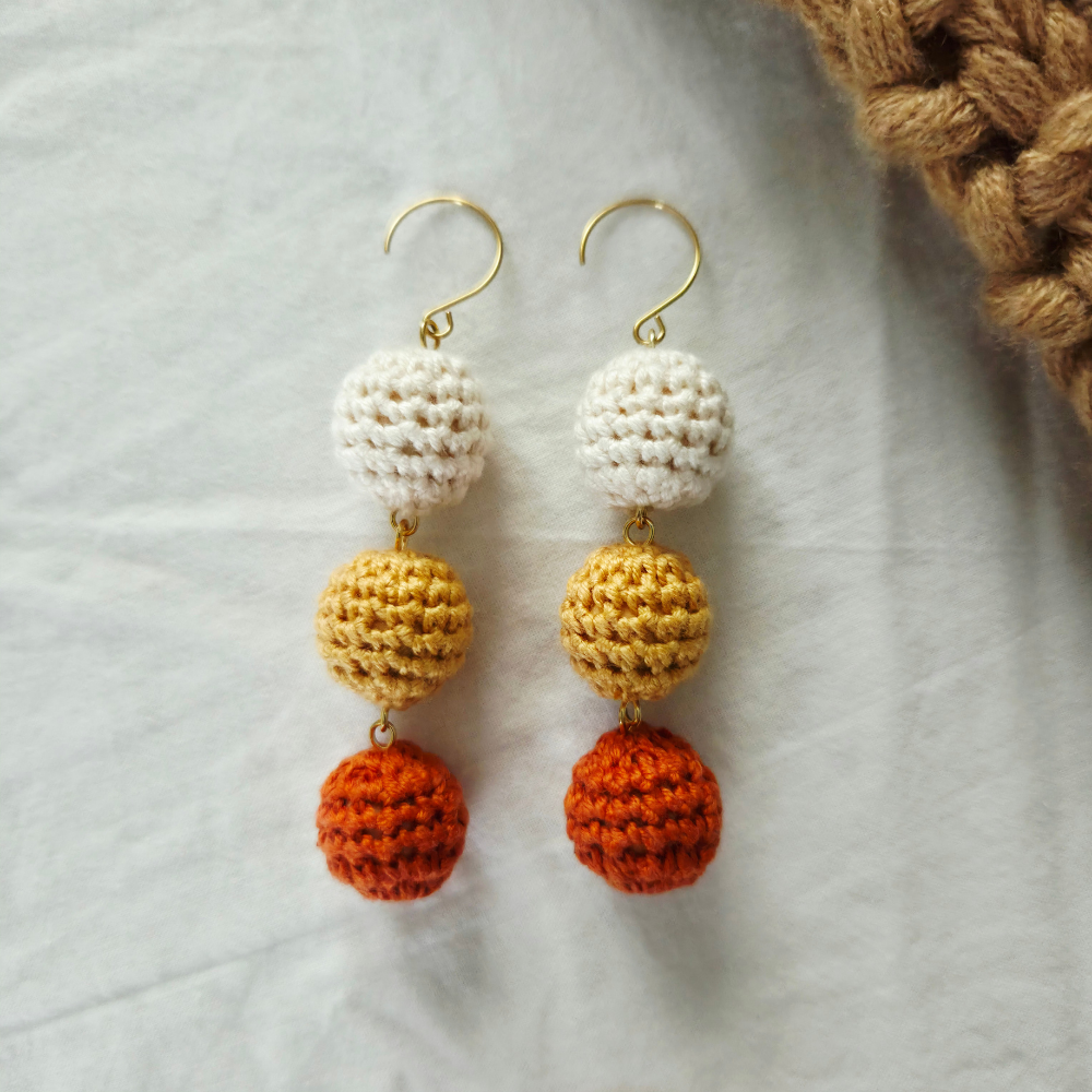 A pair of handmade crochet dangle earrings with gold-tone hooks, featuring three stacked textured crochet beads in white, mustard yellow, and burnt orange on a light gray fabric background.