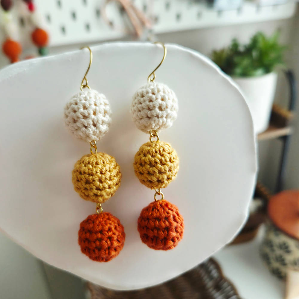 A pair of handmade crochet dangle earrings displayed on a white ceramic dish. The earrings feature three textured crochet balls in cream, mustard yellow, and burnt orange, suspended from gold-tone fishhook wires.