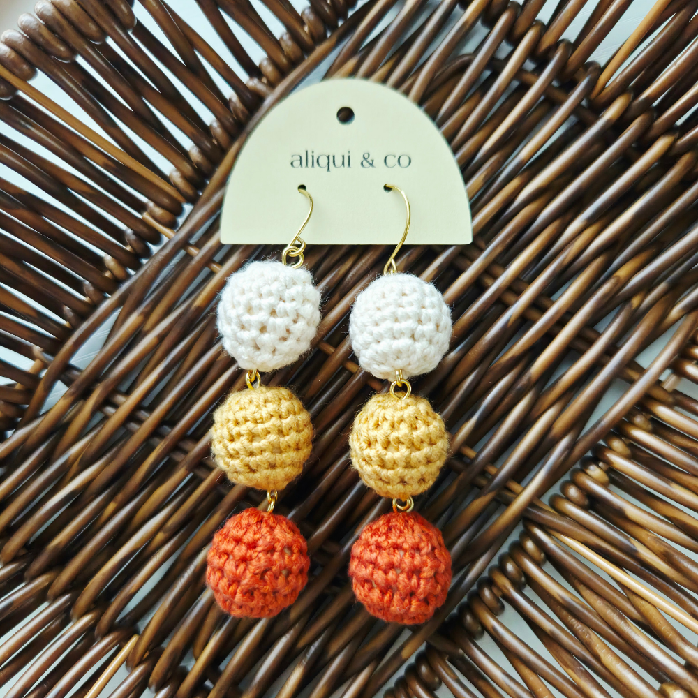 A pair of handmade crochet dangle earrings with white, yellow, and orange pom-poms on gold hooks, displayed on a cream paper tag that reads 'aliqui & co' against a brown wicker background.