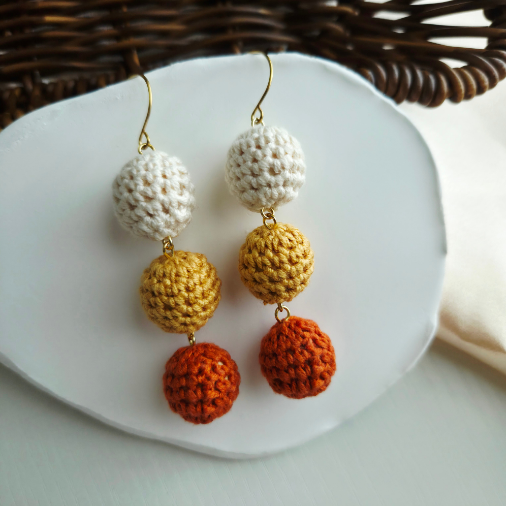 A pair of handmade crochet dangle earrings with gold fishhook wires, featuring three textured spherical drops in cream, mustard yellow, and burnt orange, displayed on a white ceramic plate.